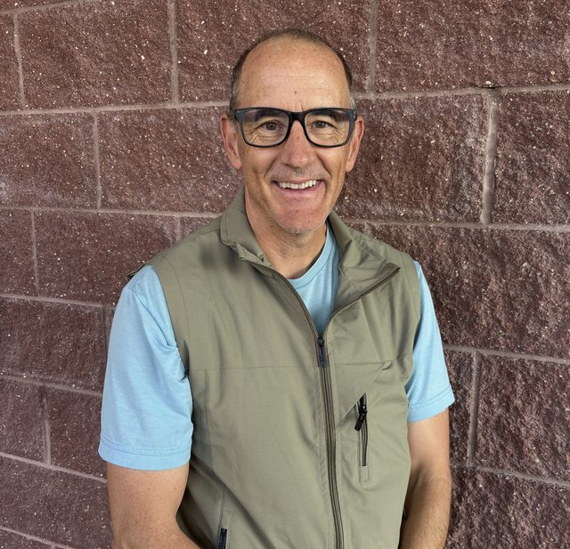 James Strawn, smiling, in a green vest and light blue T-shirt, standing against a brick wall.
